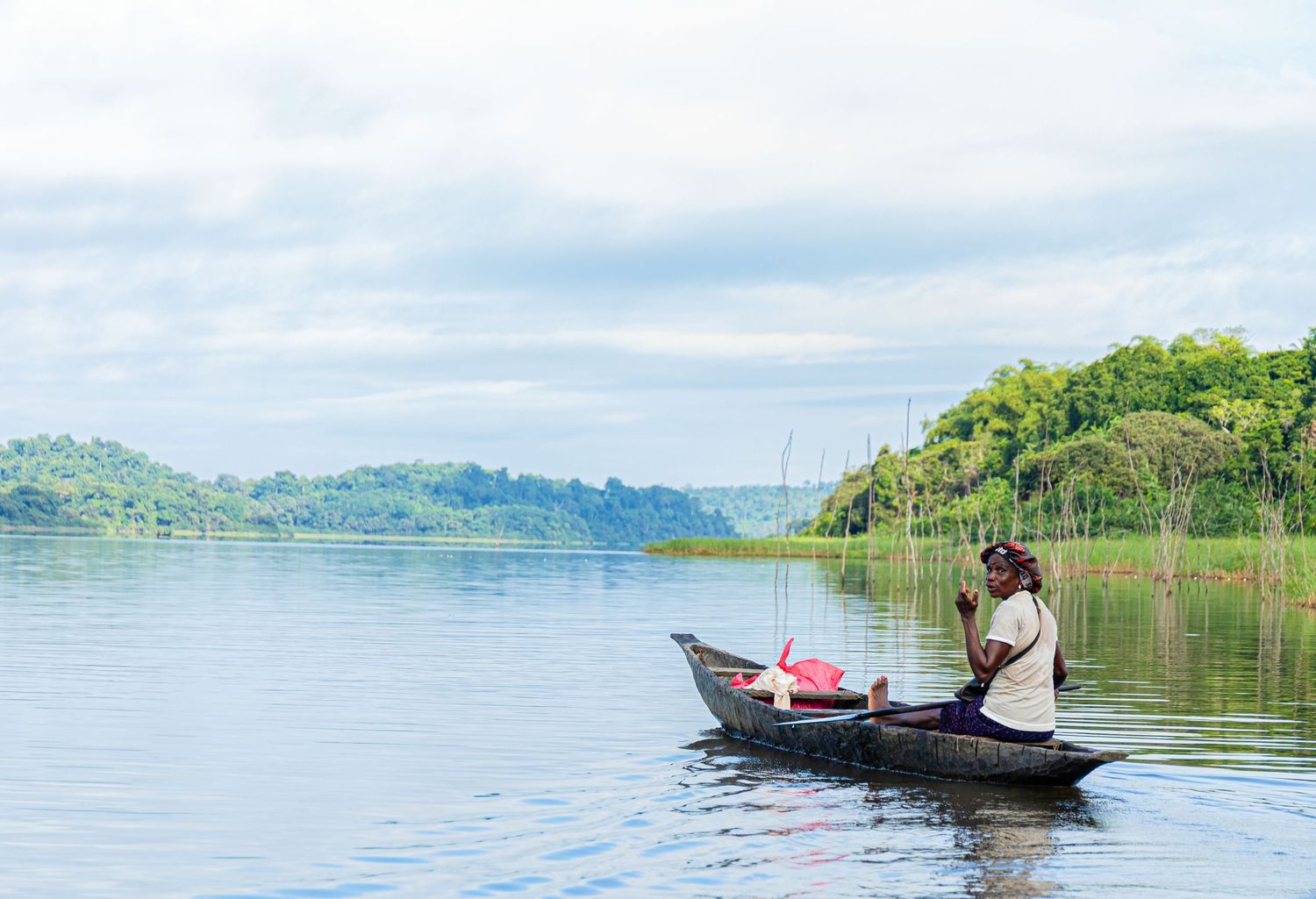 Lone fisherman sitting in a wooden canoe on a calm lake surrounded by lush green hills.