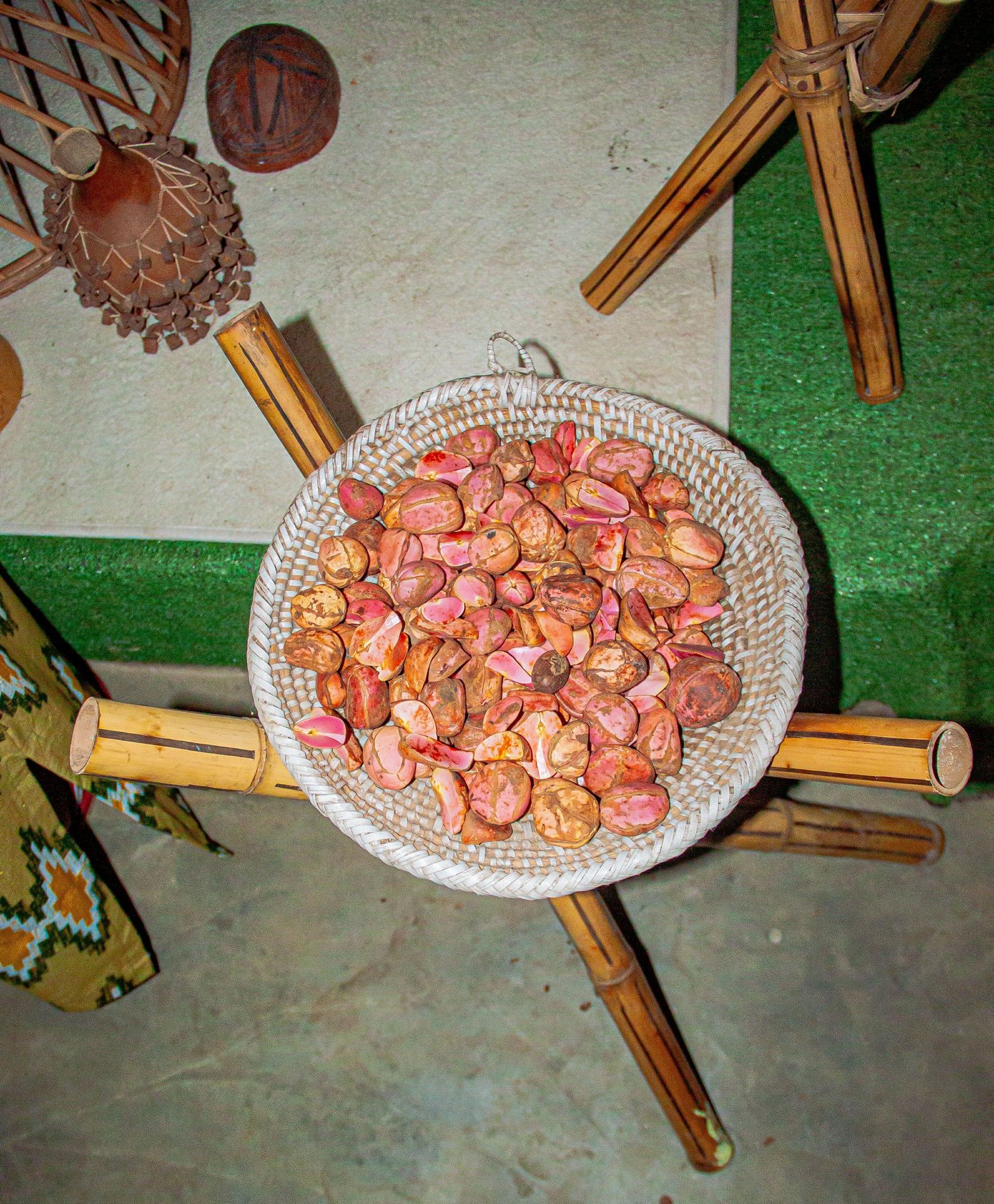 Red kola nuts displayed in a woven basket on a bamboo stand surrounded by traditional African decor.