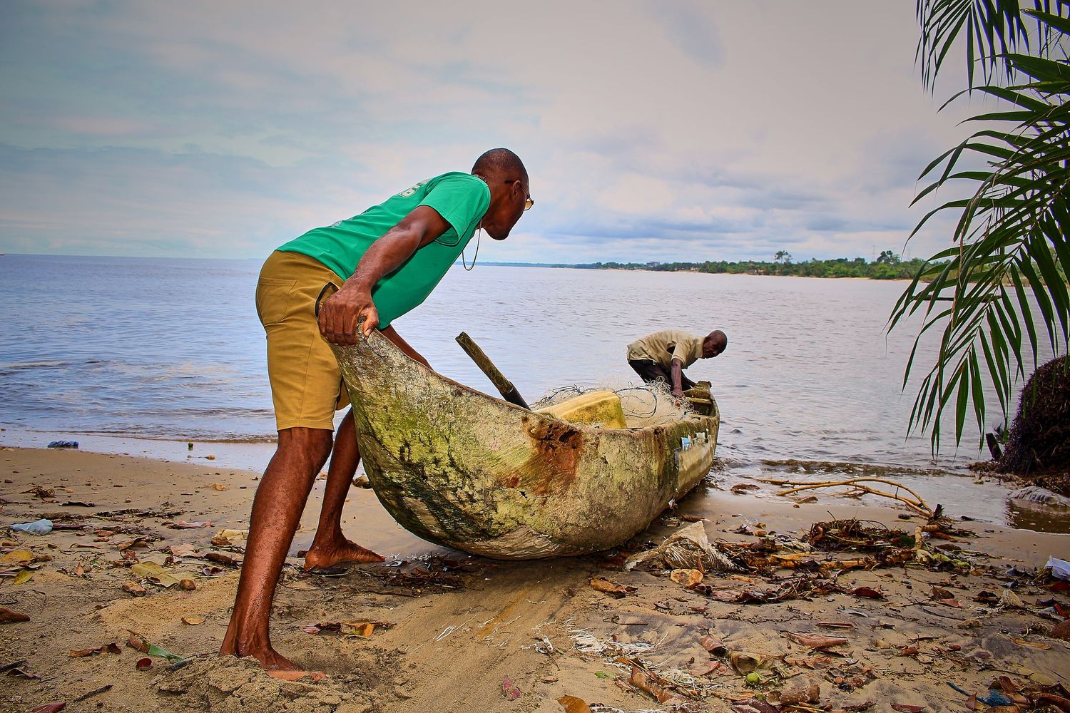 Man pushing a weathered dugout canoe on a tropical sandy beach near calm waters.