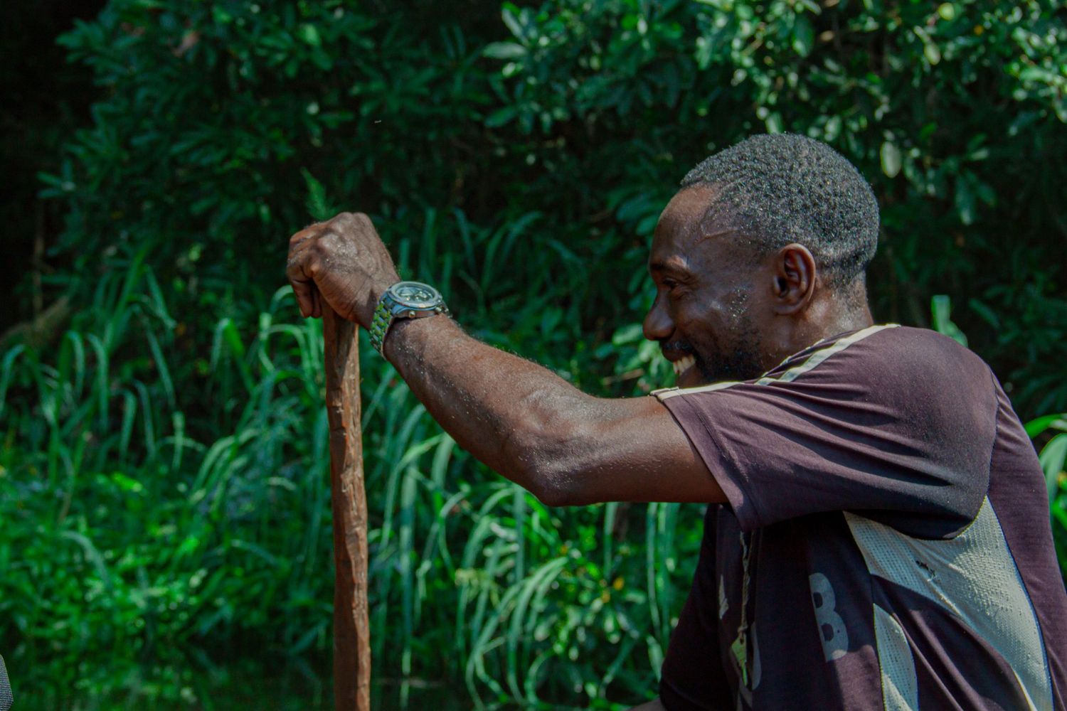 Man checking watch while leaning on a wooden stick in a green forest.