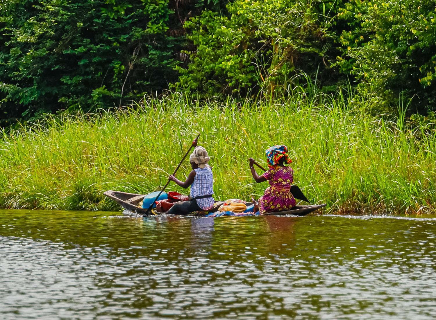 Two people paddling a wooden canoe along a lush green riverbank in tropical Africa.