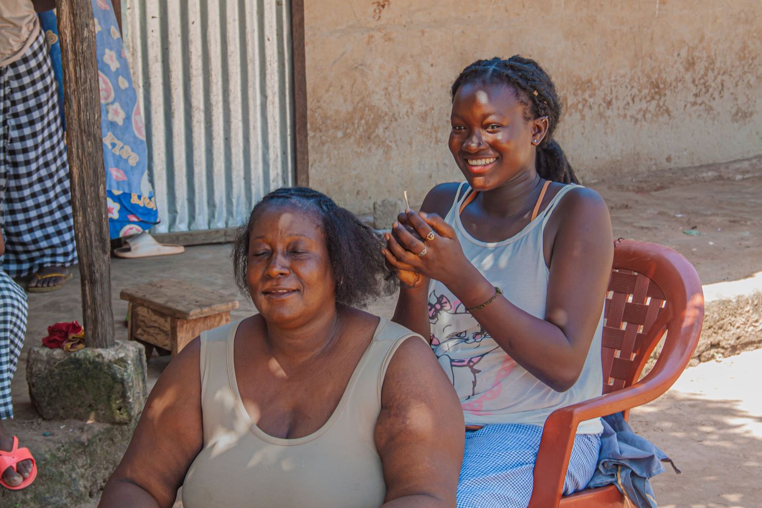 Two African women smiling outdoors, one braiding the others hair.