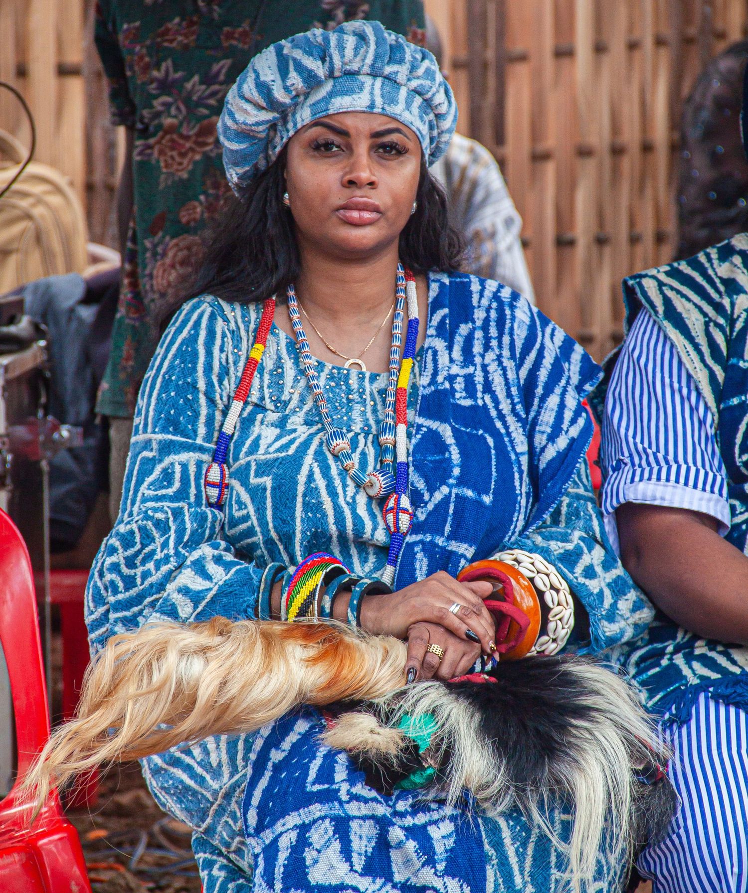 Young African woman in blue traditional dress with colorful beaded necklaces holding decorative tail at cultural event.