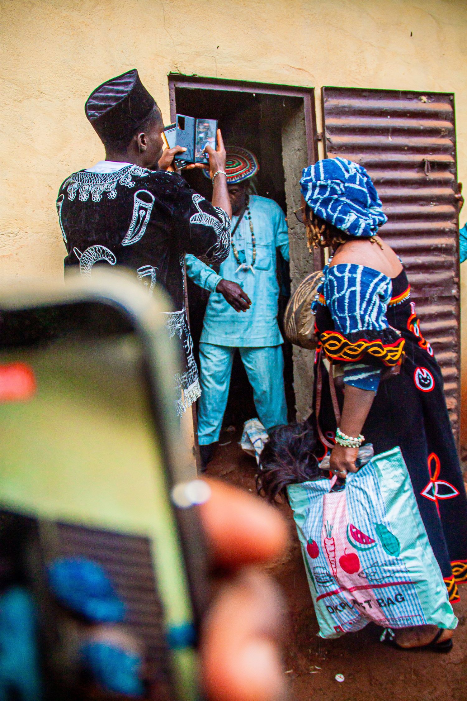 People in traditional African attire gather at a doorway, with one person photographing the moment on a smartphone.