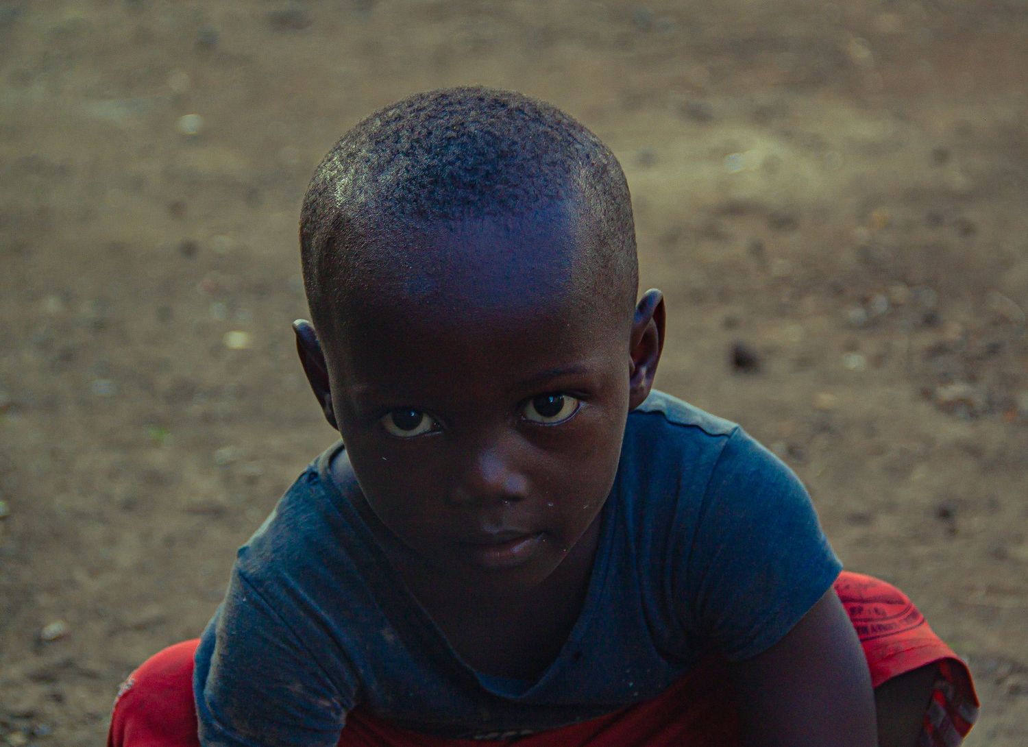 A close-up portrait of a young African boy in a blue shirt looking directly at the camera.