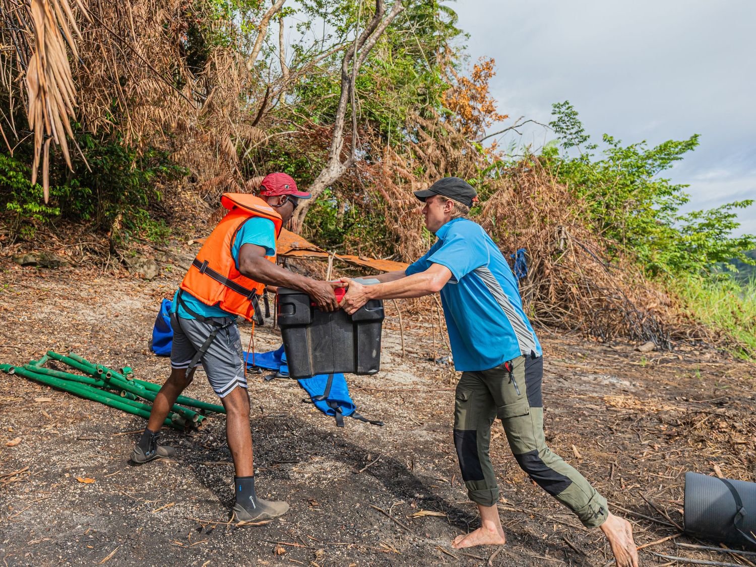 Two workers in orange vests carry heavy black containers during an outdoor conservation cleanup effort near tropical vegetation.
