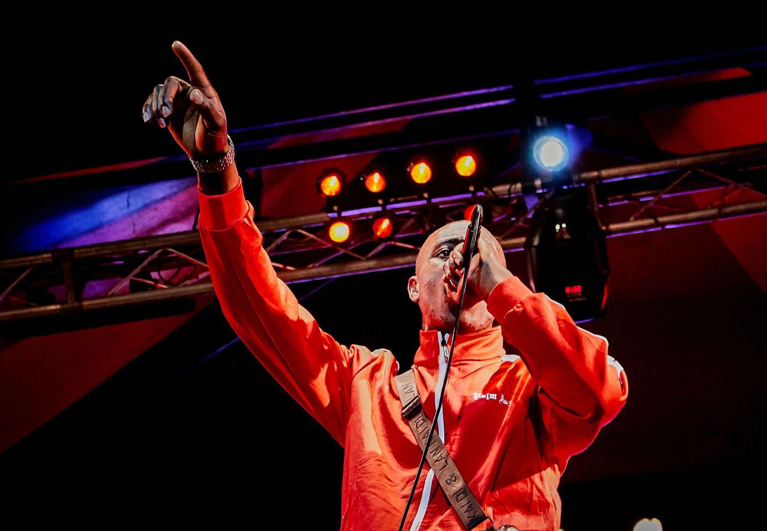 Energetic performer in an orange jacket pointing upward while singing into a microphone on a lit concert stage.