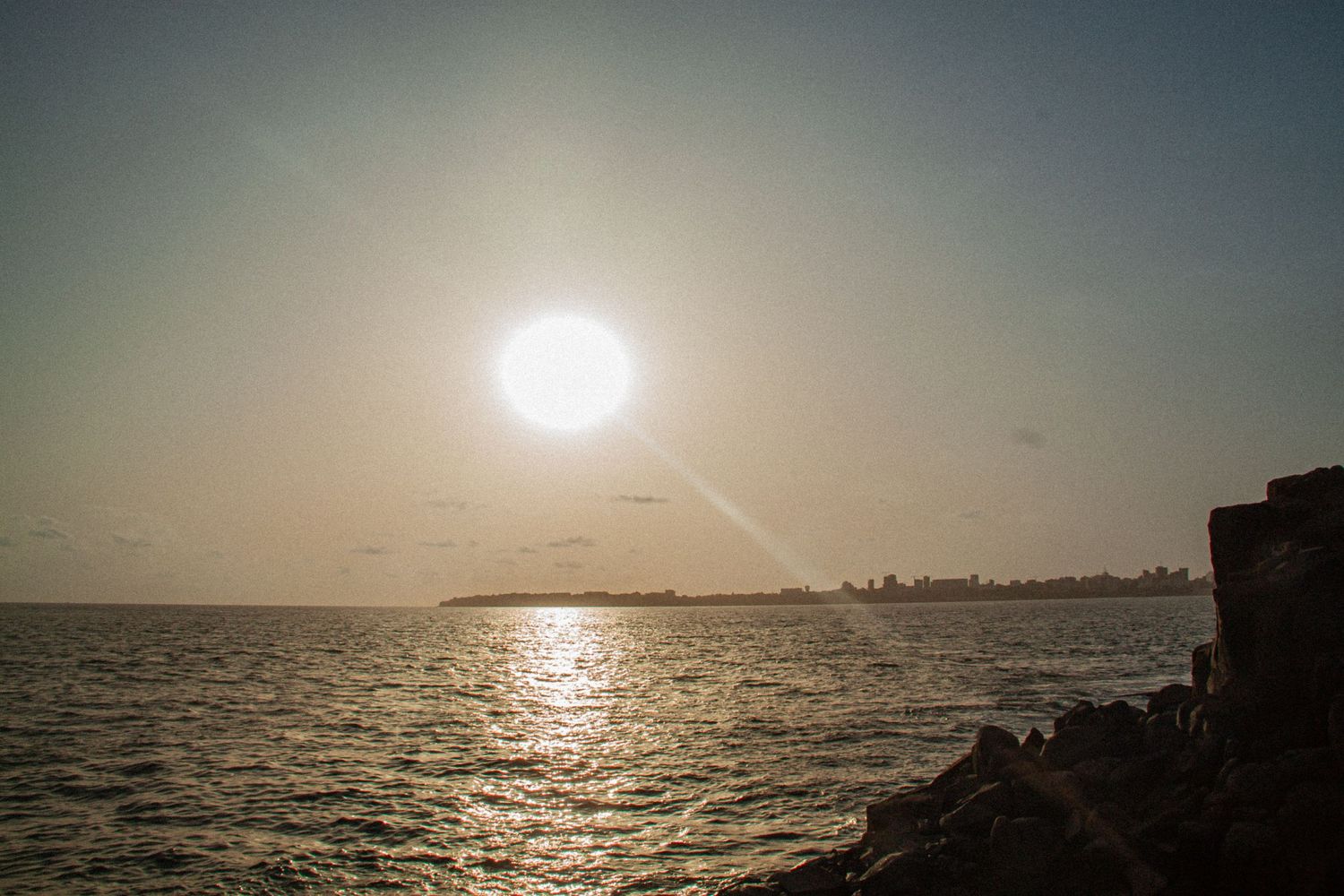 Golden sunset reflecting on calm ocean waters viewed from rocky shoreline with distant city skyline on the horizon.