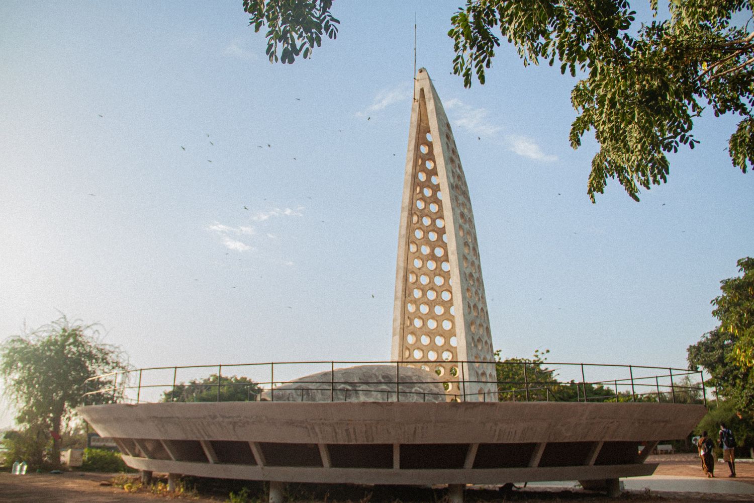 Modern latticed tower monument rising above circular platform surrounded by trees at dusk.