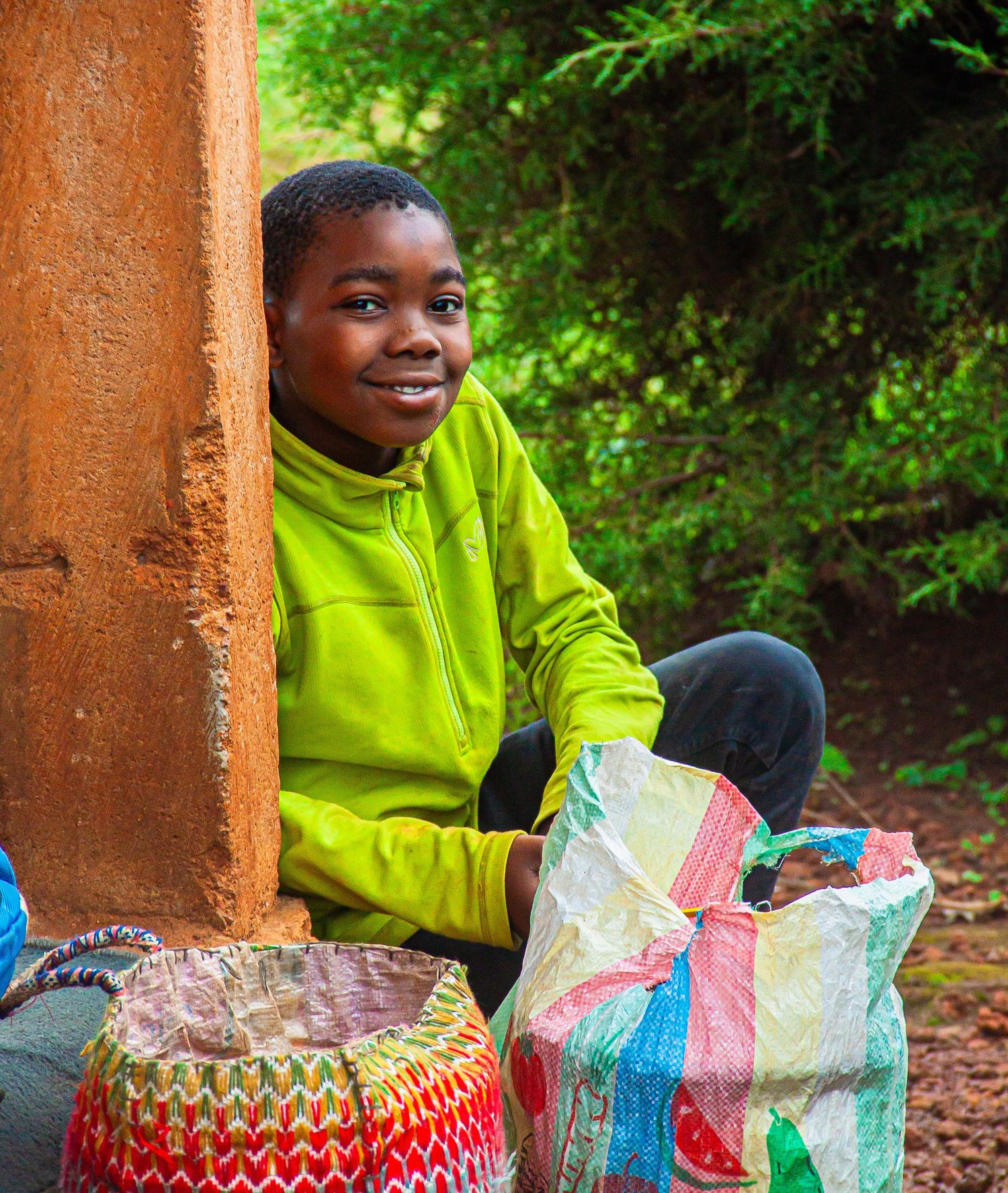 Smiling African boy in green hoodie sitting outdoors with colorful woven baskets and bags.