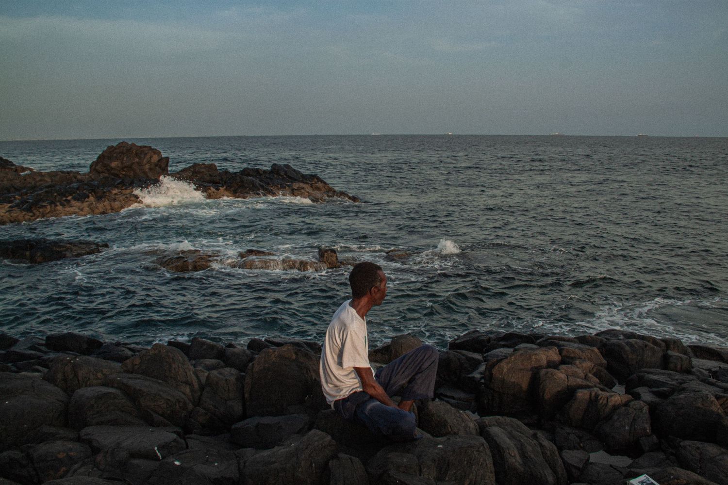 A man sits alone on rocky coastal cliffs gazing at ocean waves crashing against rocks at dusk.