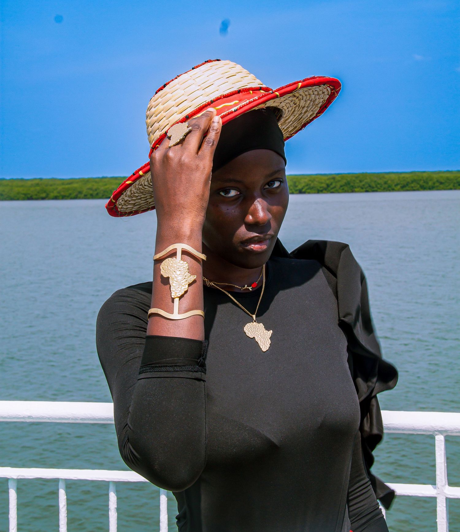 Young African woman in black outfit wearing straw hat with red trim and gold Africa map jewelry by waterfront.