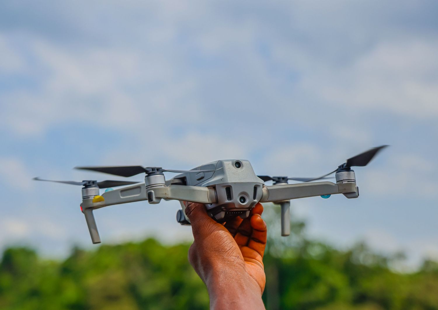 Hand holding a DJI drone ready for launch against a cloudy blue sky with green trees.