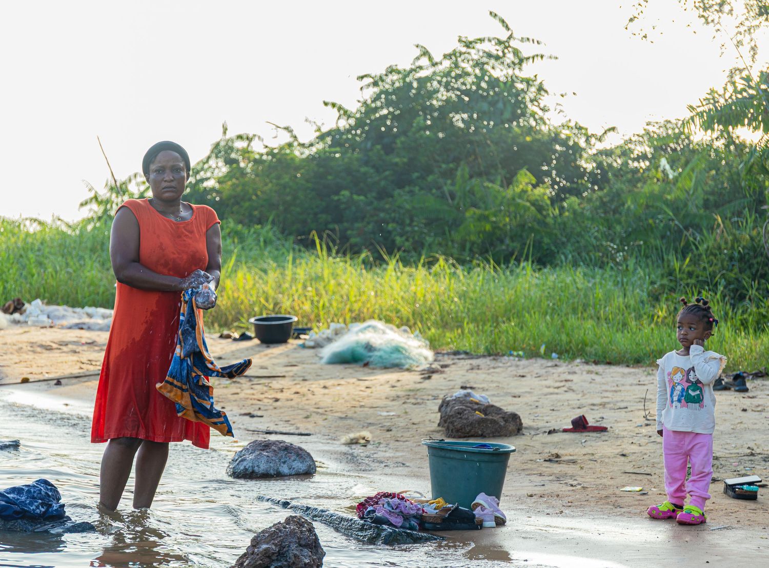African woman in red dress doing laundry on a sandy riverbank with a young child nearby.