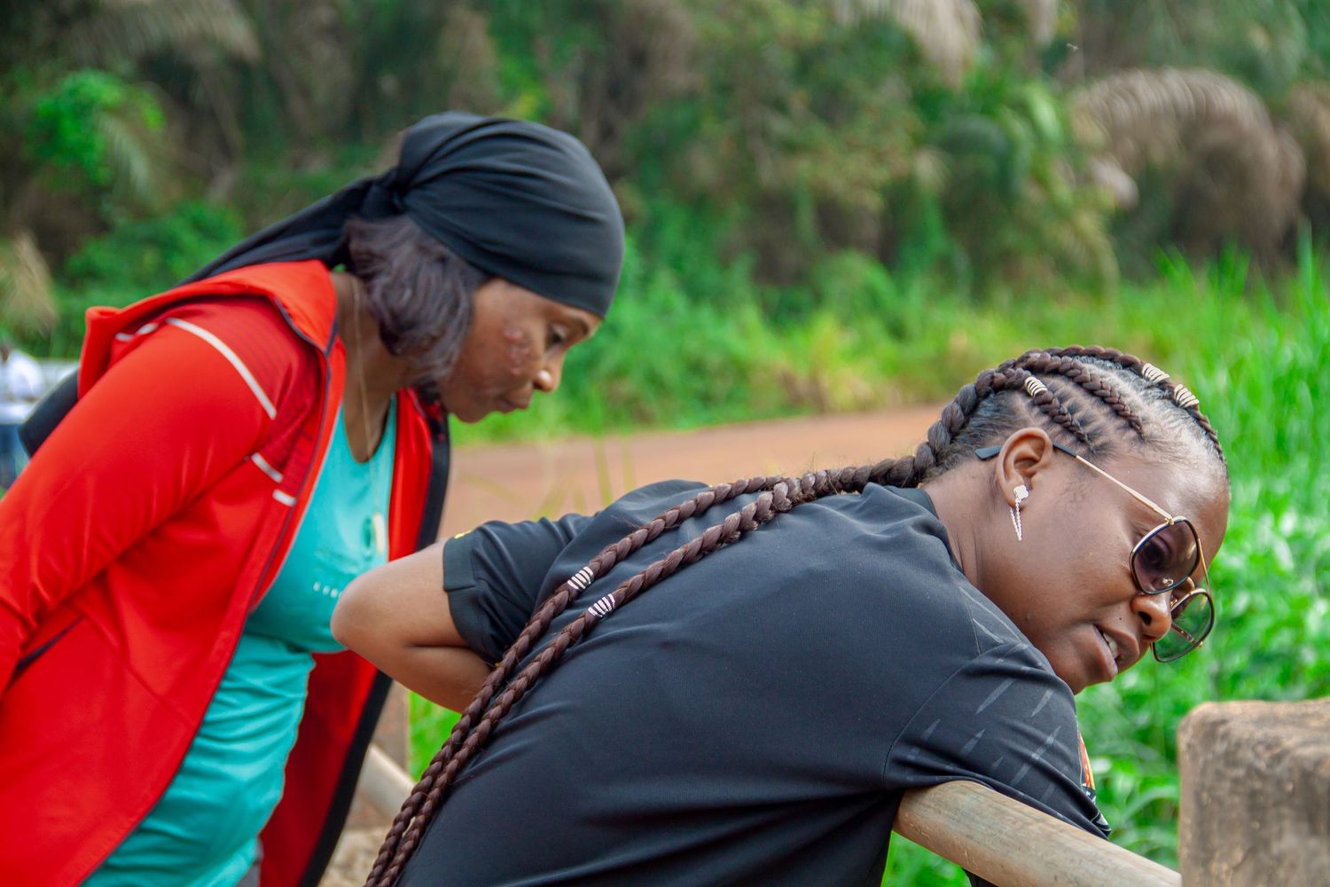 Two African women leaning on a wooden railing outdoors surrounded by green vegetation in Cameroon.