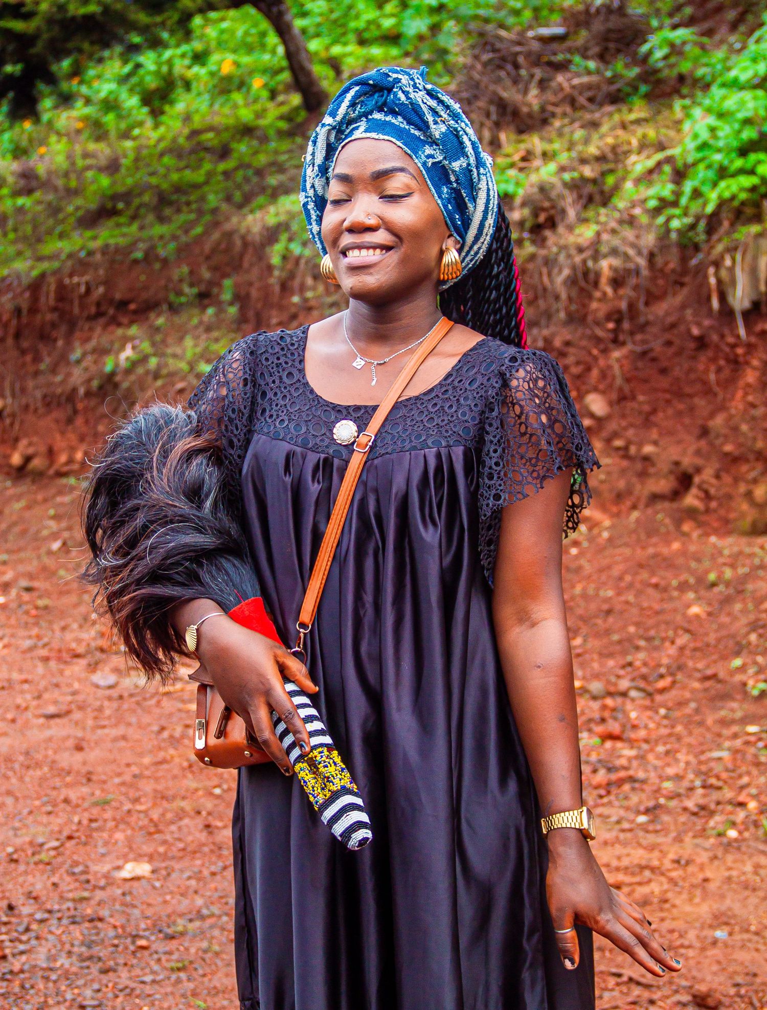 Smiling African woman in black dress and blue head wrap holding a feather duster outdoors.