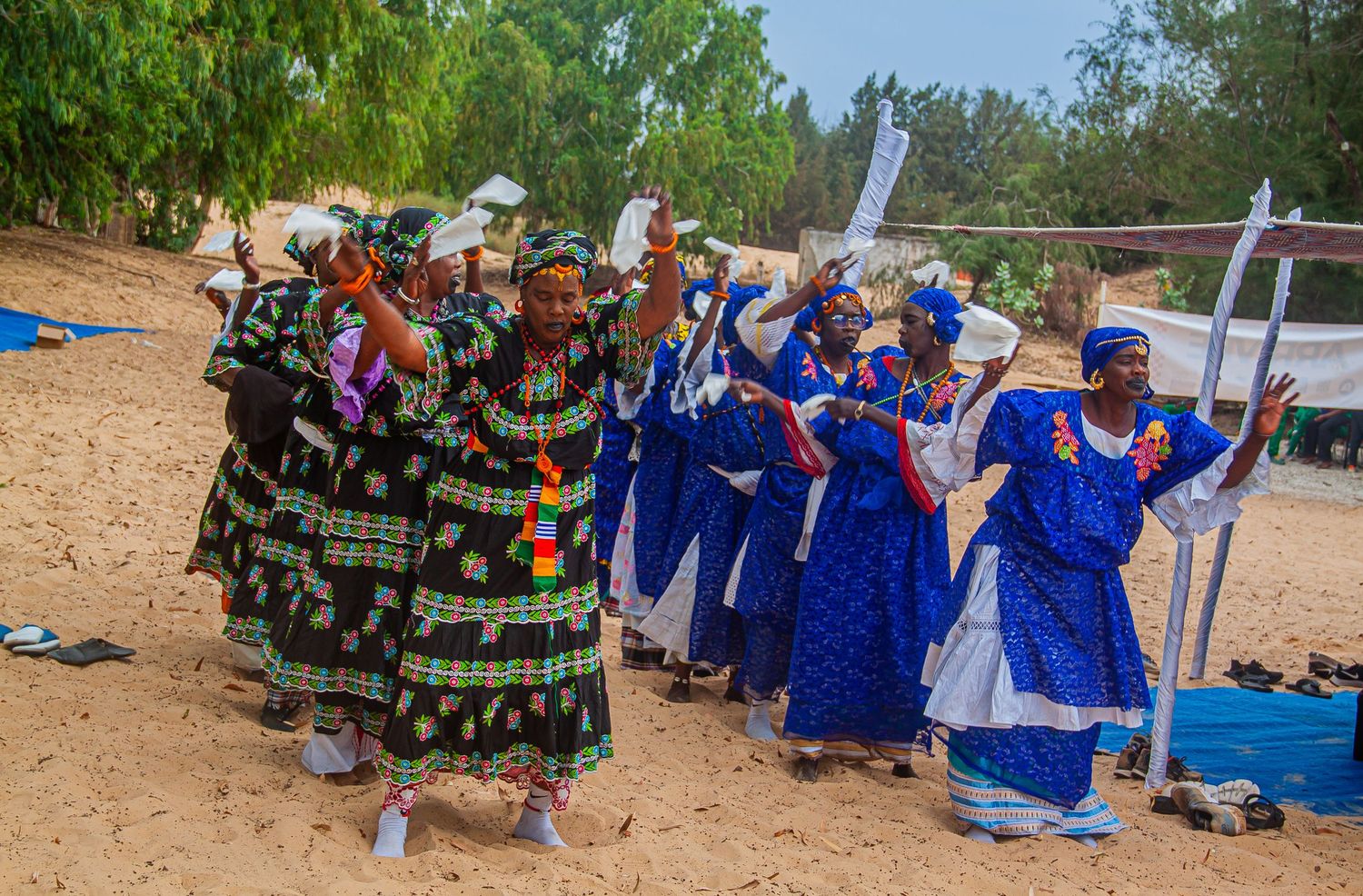 Women in vibrant traditional African attire dance joyfully outdoors on sandy ground during a cultural celebration.