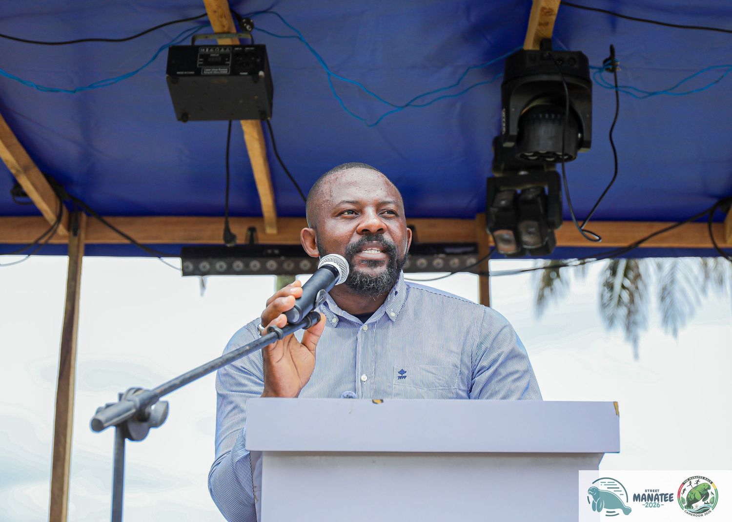A man in a blue shirt delivers a speech at an outdoor podium with a microphone.
