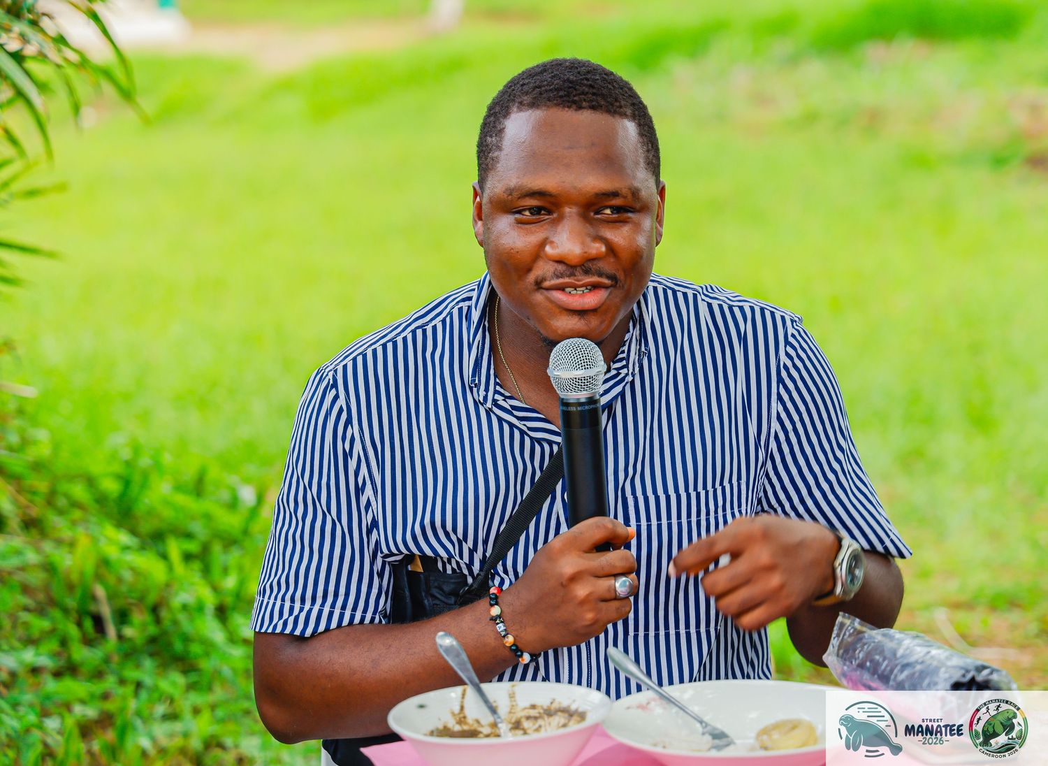 Smiling man in striped shirt speaking into microphone while eating outdoors.
