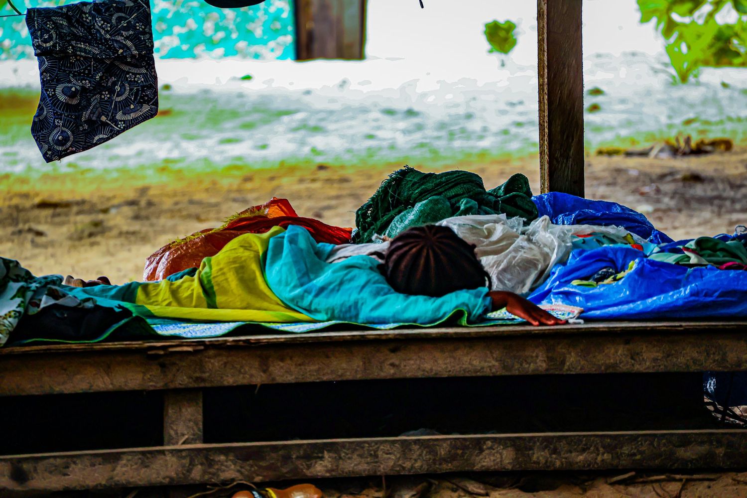 Colorful folded clothes and fabrics piled on a wooden bench under an open shelter.