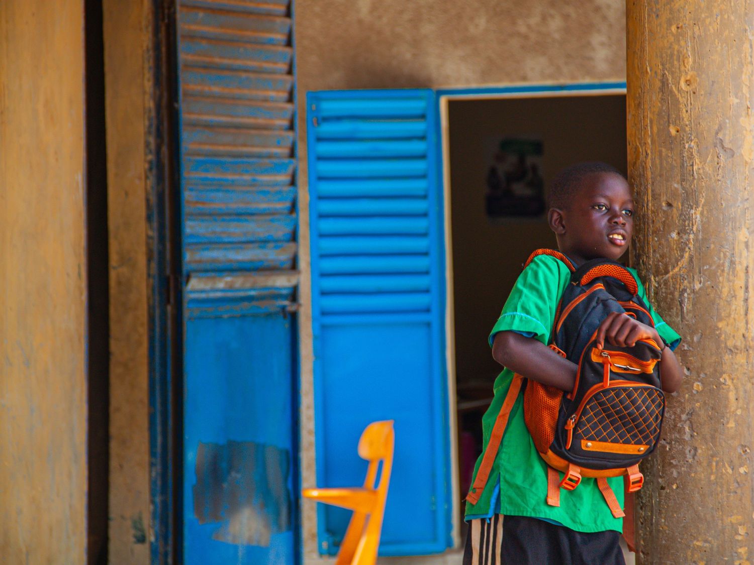 Young African boy with backpack standing by a colorful blue door in a school building.
