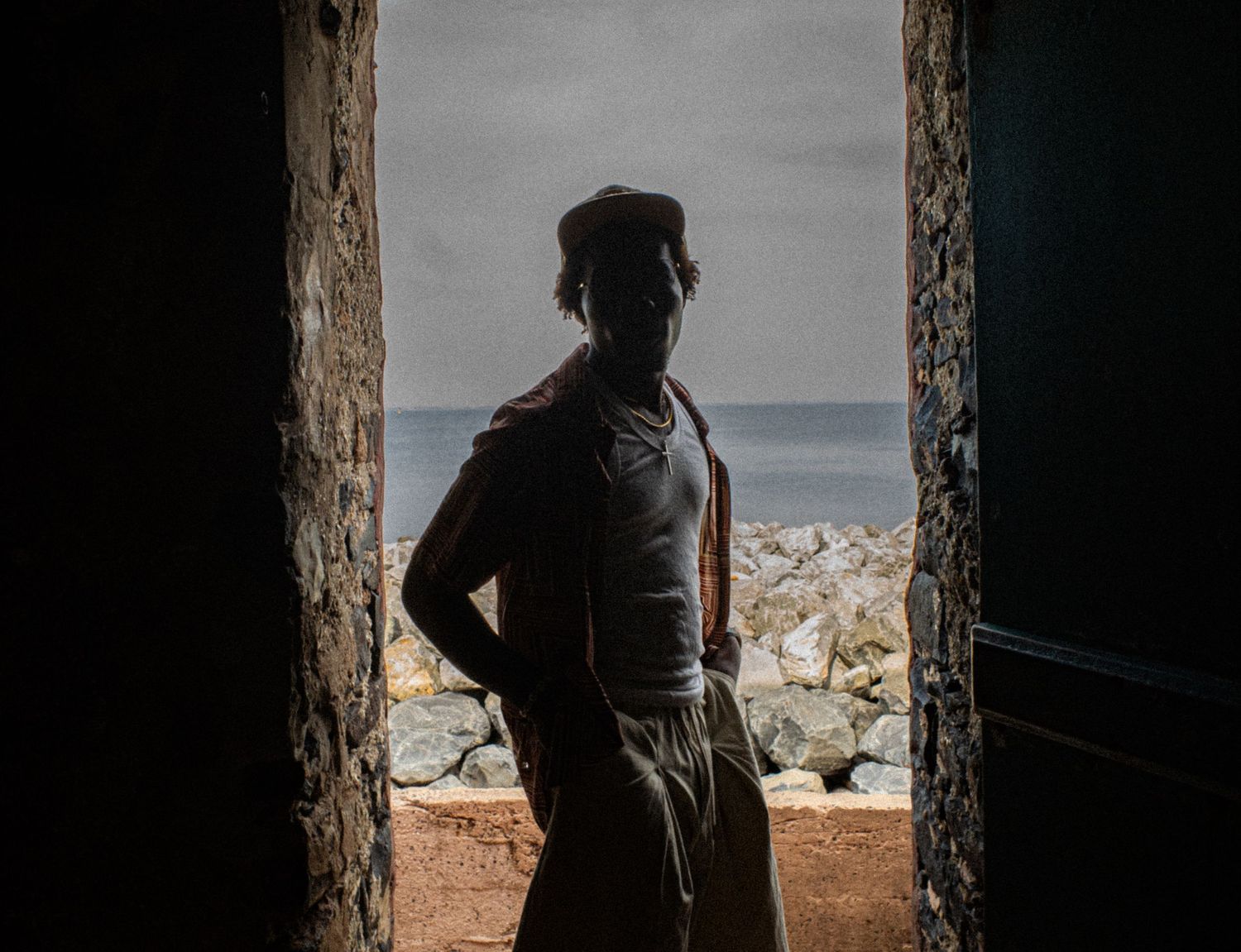 A silhouetted figure stands in a doorway overlooking rocky shores and the ocean in soft dramatic light.