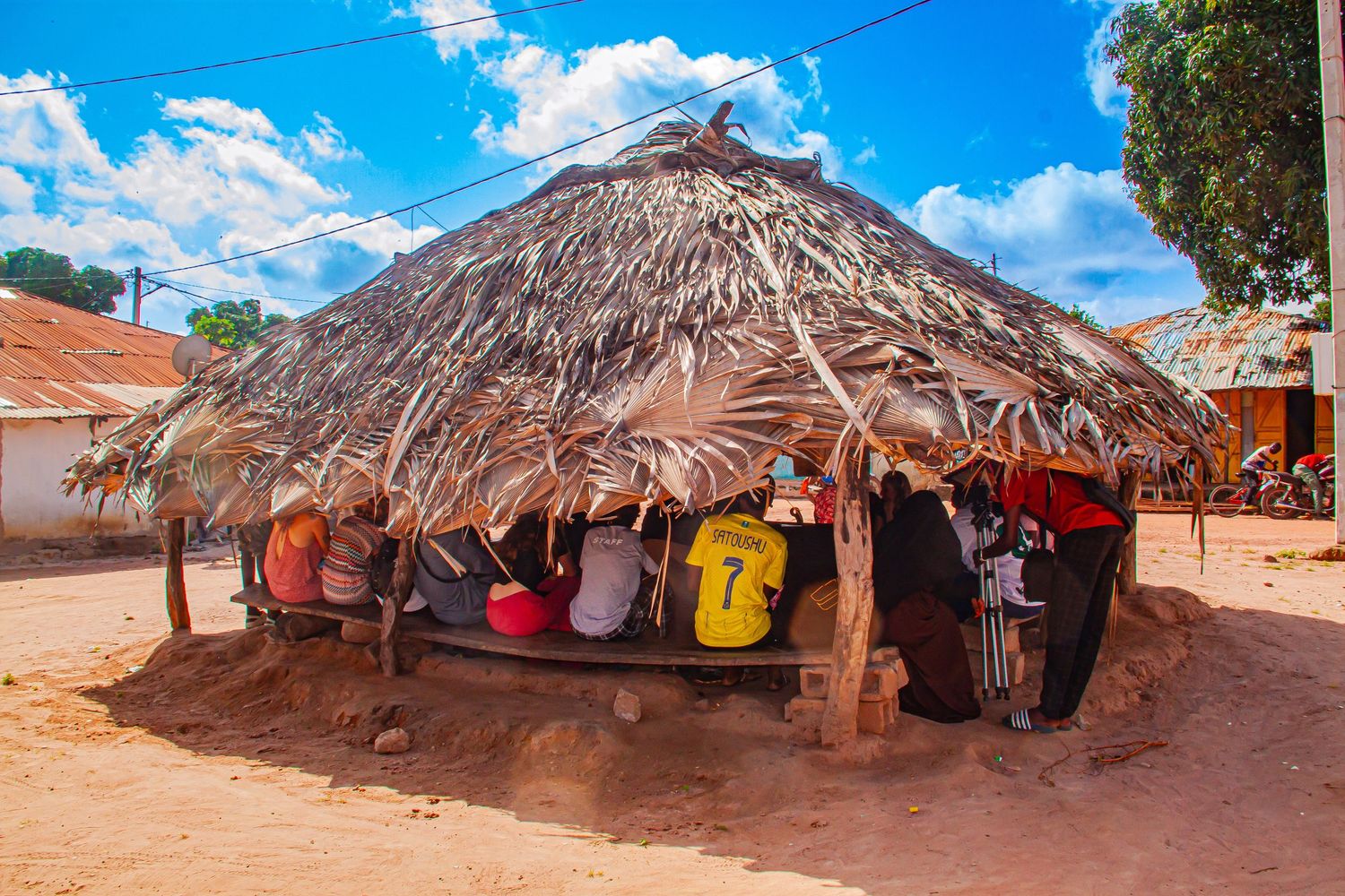 People gathered under a traditional thatched roof hut in an African village on a sunny day.