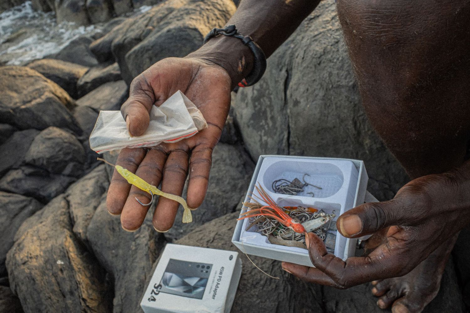 Close-up of hands holding fishing lures and tackle on rocky shoreline in Senegal.