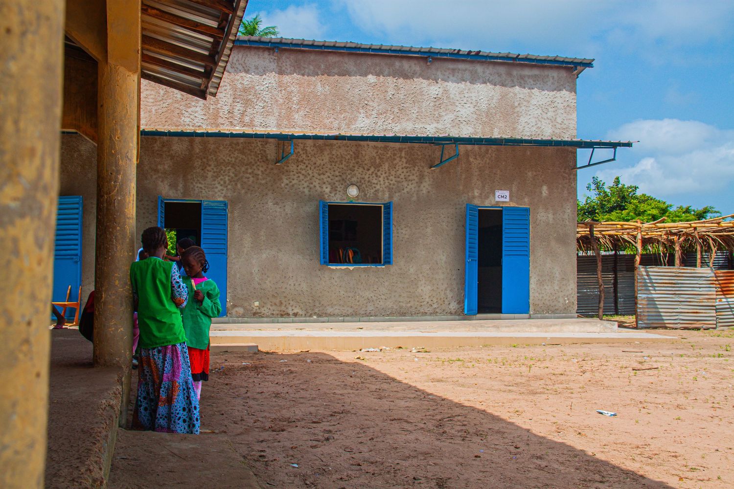 Small grey concrete building with blue doors and windows on a dusty courtyard in rural Africa.