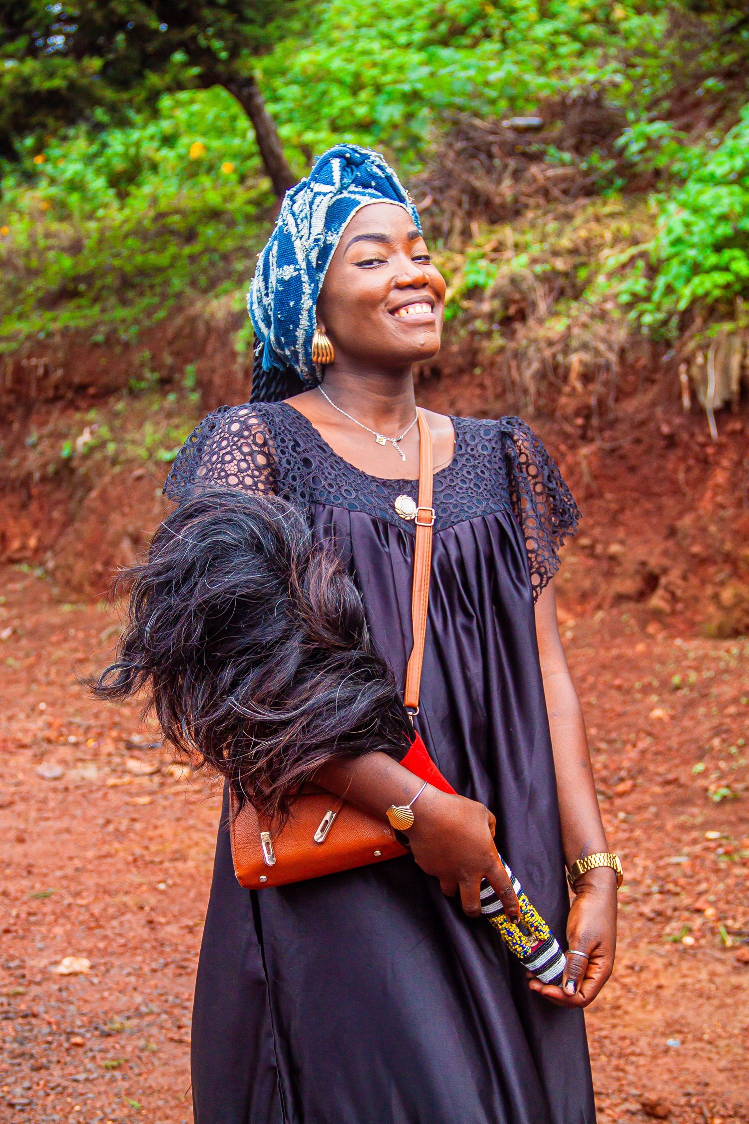 Smiling African woman in traditional blue head wrap and black dress holding a feathered bag outdoors.