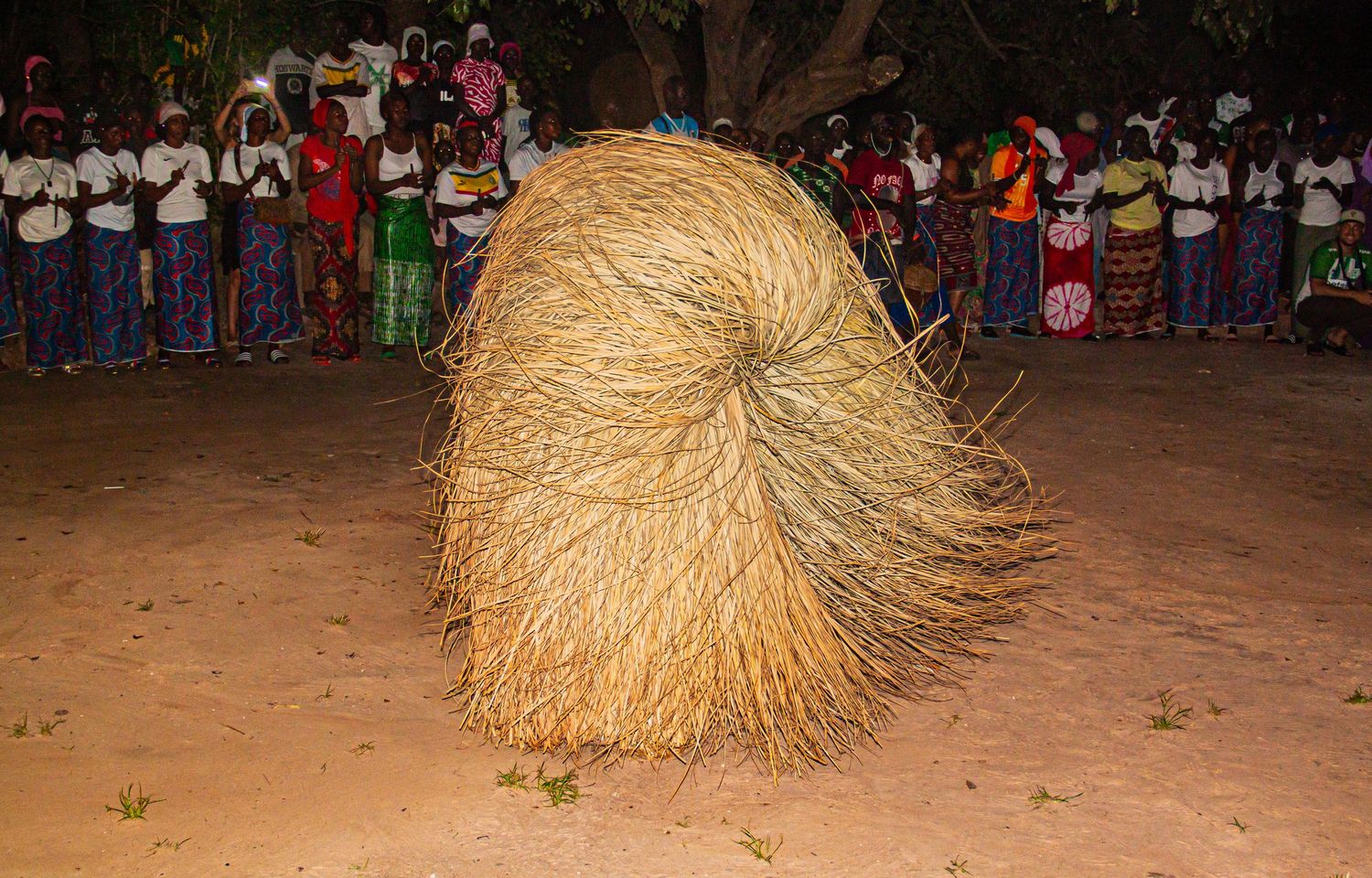 A performer covered in a large grass costume dances at a traditional African night ceremony with onlookers.