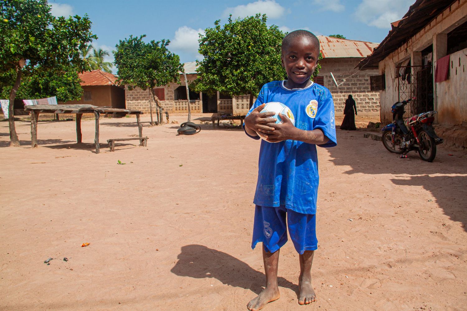 African child in a blue jersey holding a soccer ball while standing barefoot on dry dirt in a rural village.