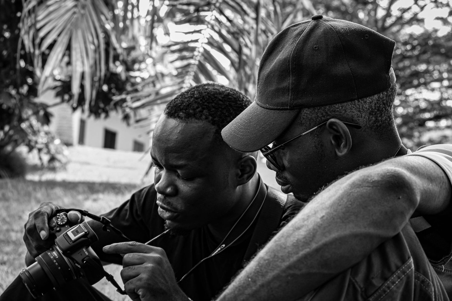 Black and white photo of two African men reviewing photos on a camera outdoors.