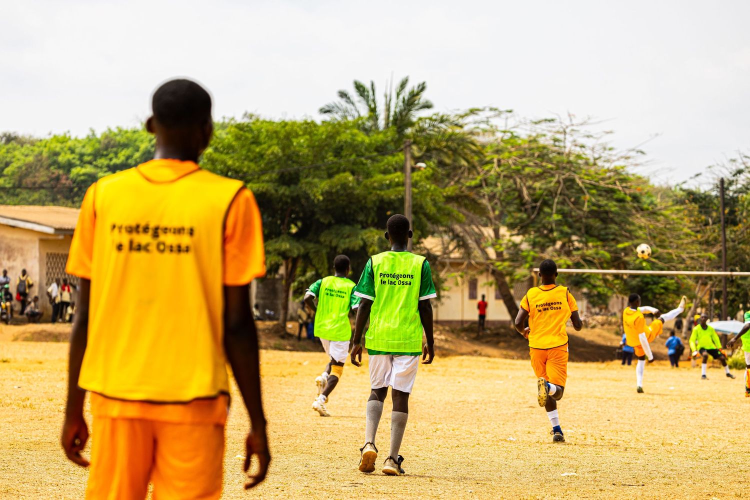 Children in yellow and green jerseys play soccer on a dirt field with trees in the background.