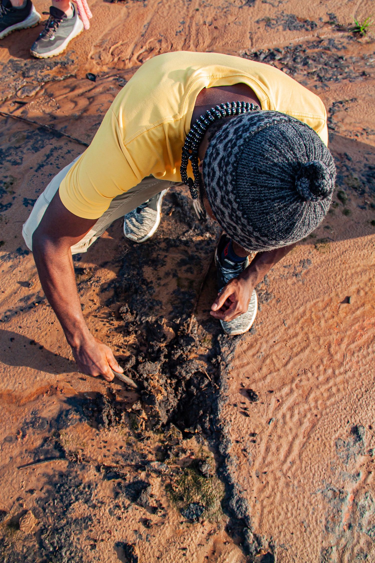 Person in yellow shirt and knit hat crouching to examine dark soil on red sand desert ground.