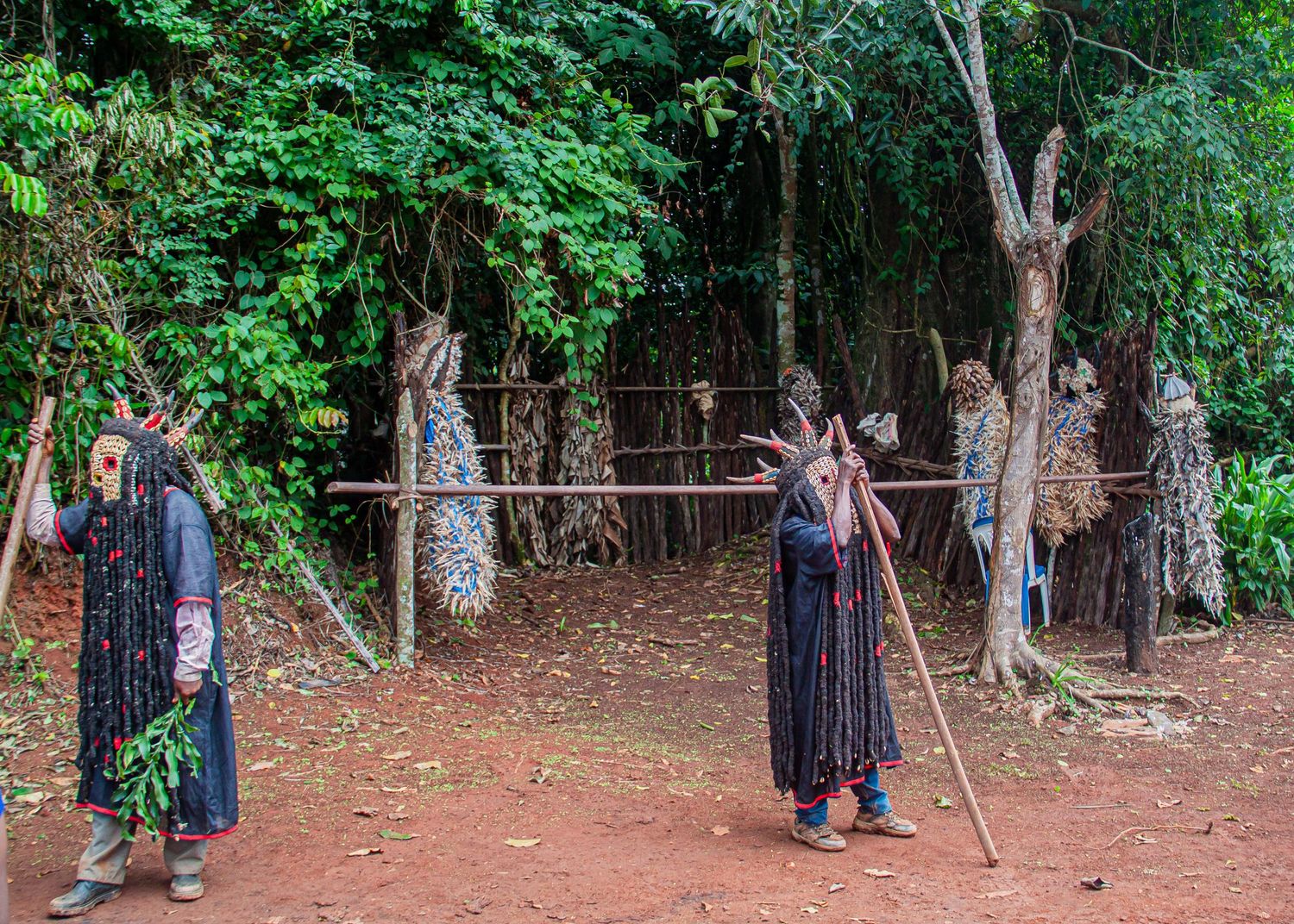 Traditional African masked figures in grass costumes standing on red earth near forest trees.