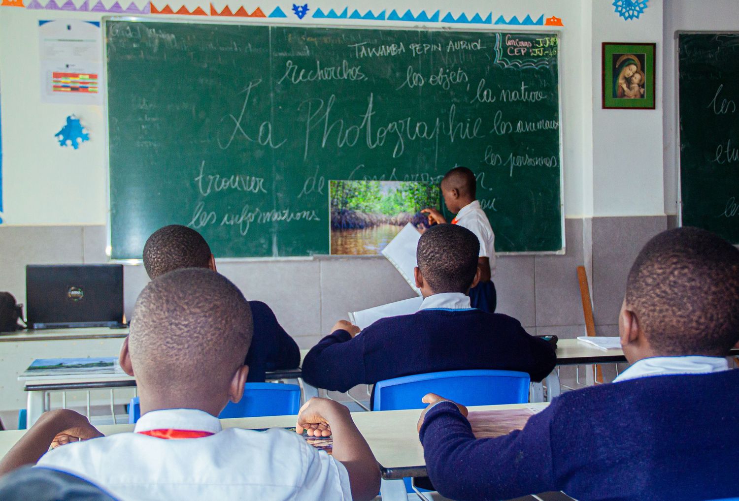 A teacher writes French lesson on a green chalkboard while students sit at desks in an African classroom.