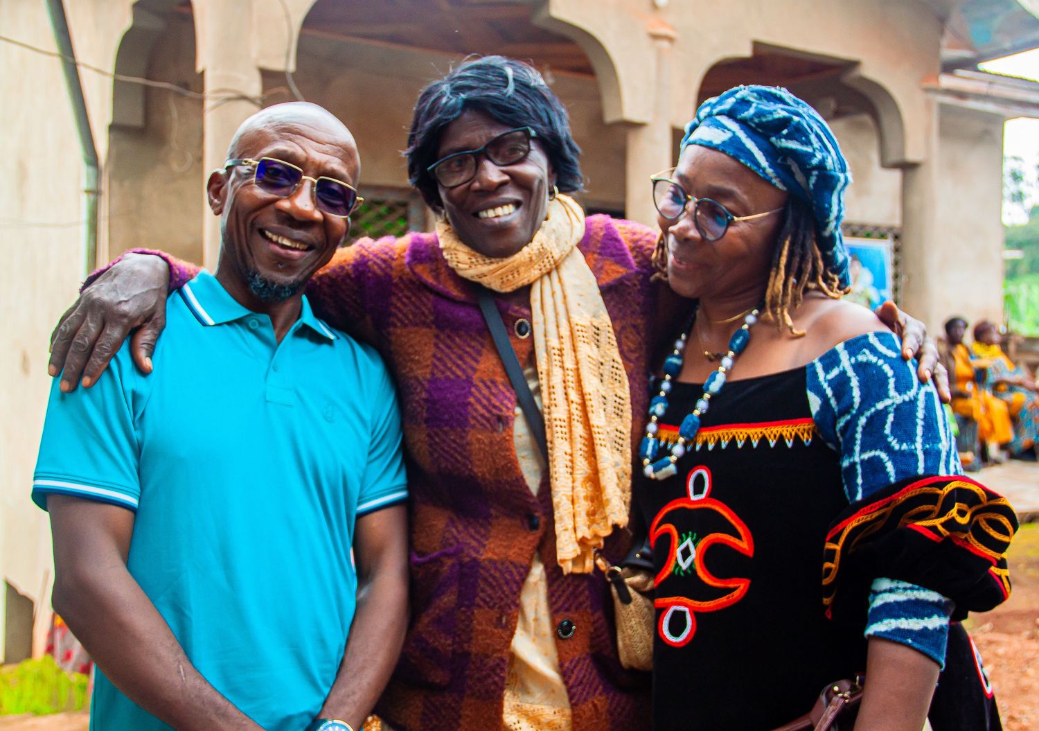 Three smiling people in colorful traditional African attire pose together outdoors at a festive gathering.