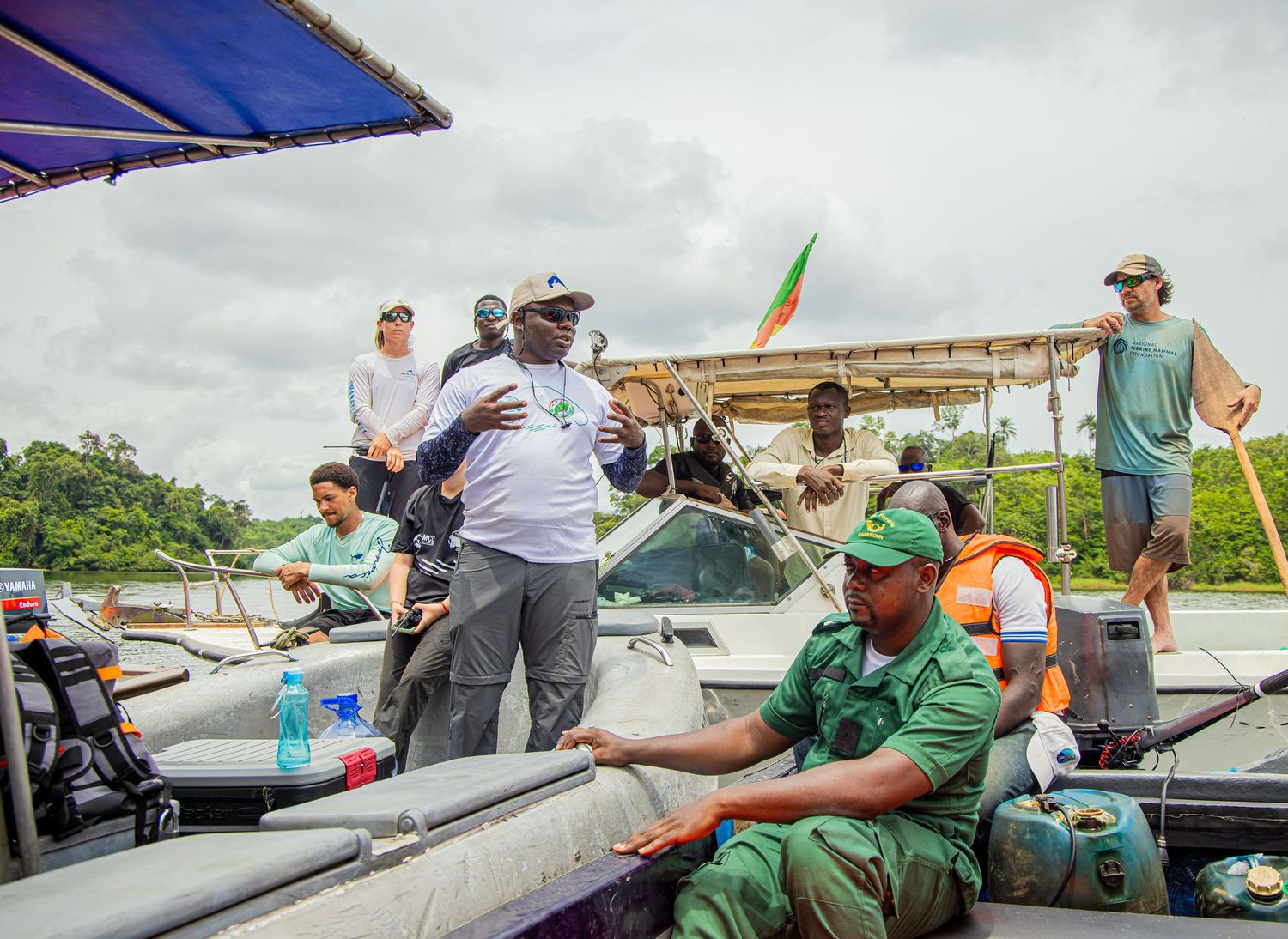 A man in a white shirt gestures while speaking to a group on boats during a river conservation expedition in Africa.