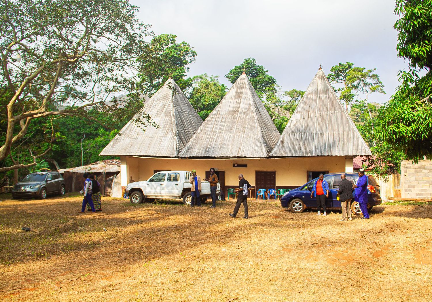 Traditional African village with thatched roof huts, white pickup truck, and locals gathered on red dirt ground.