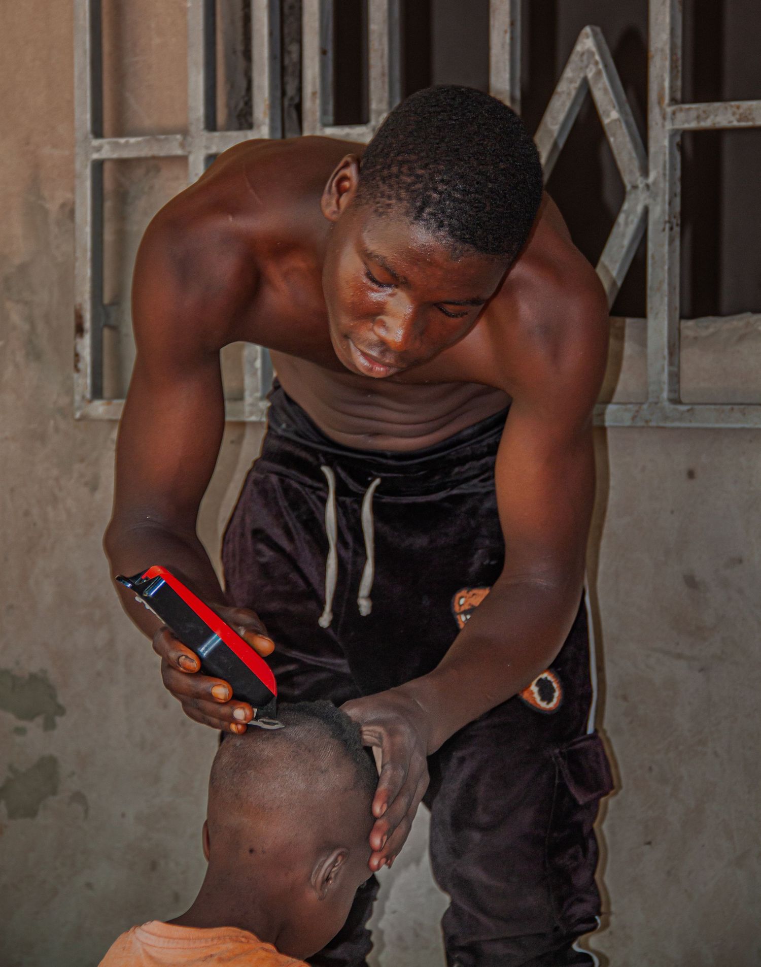 Shirtless young man giving a haircut outdoors using electric clippers in an African urban setting.