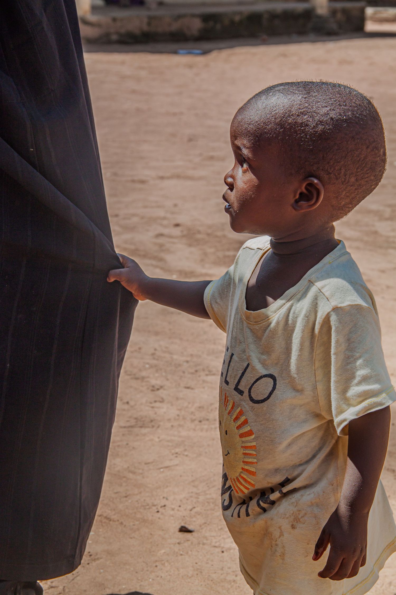 A small African toddler in a Hello shirt leans against a dark fabric in bright sunlight.
