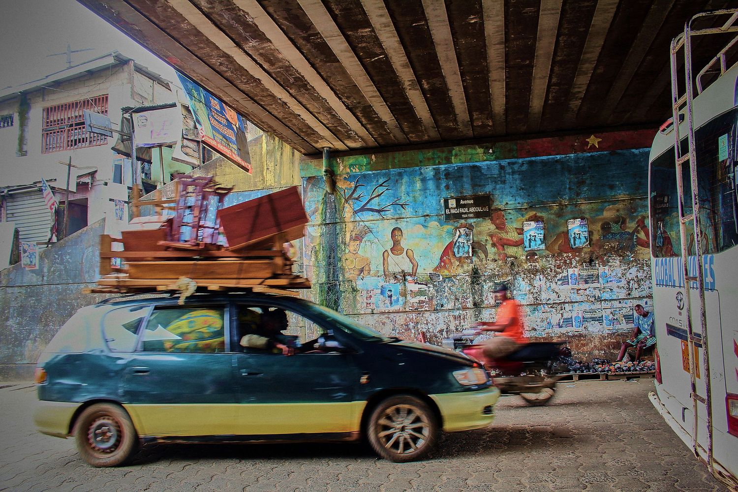 Overloaded car carrying wooden furniture drives under a colorful urban mural in an African city.