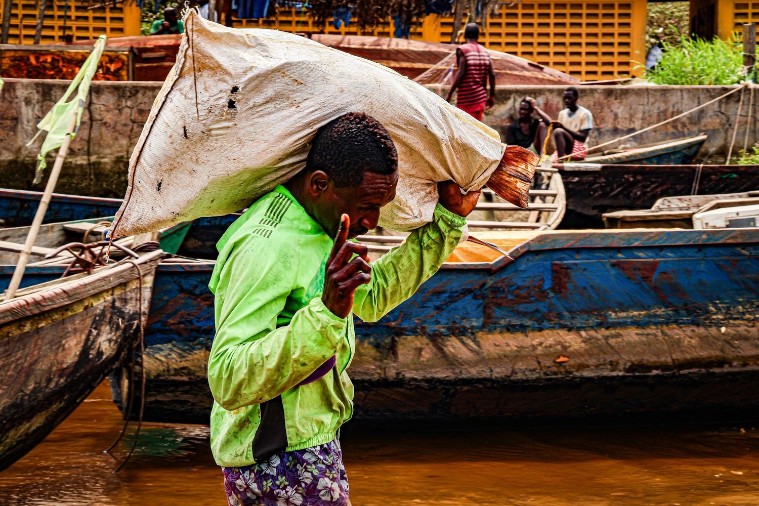 A man in a green shirt carries a large sack on his shoulder near colorful wooden boats on a river dock.