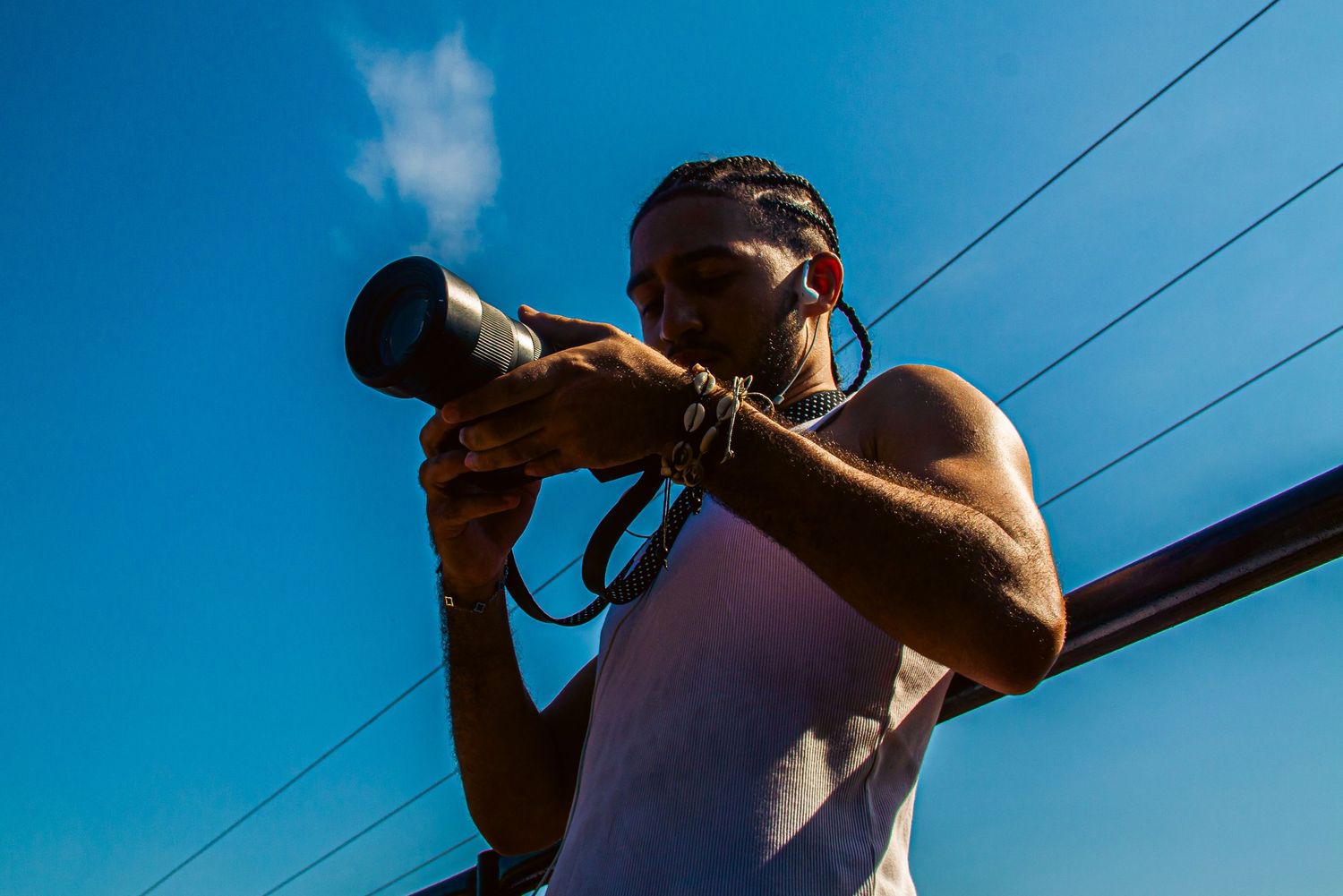 Man in white tank top photographing with telephoto lens against bright blue sky.