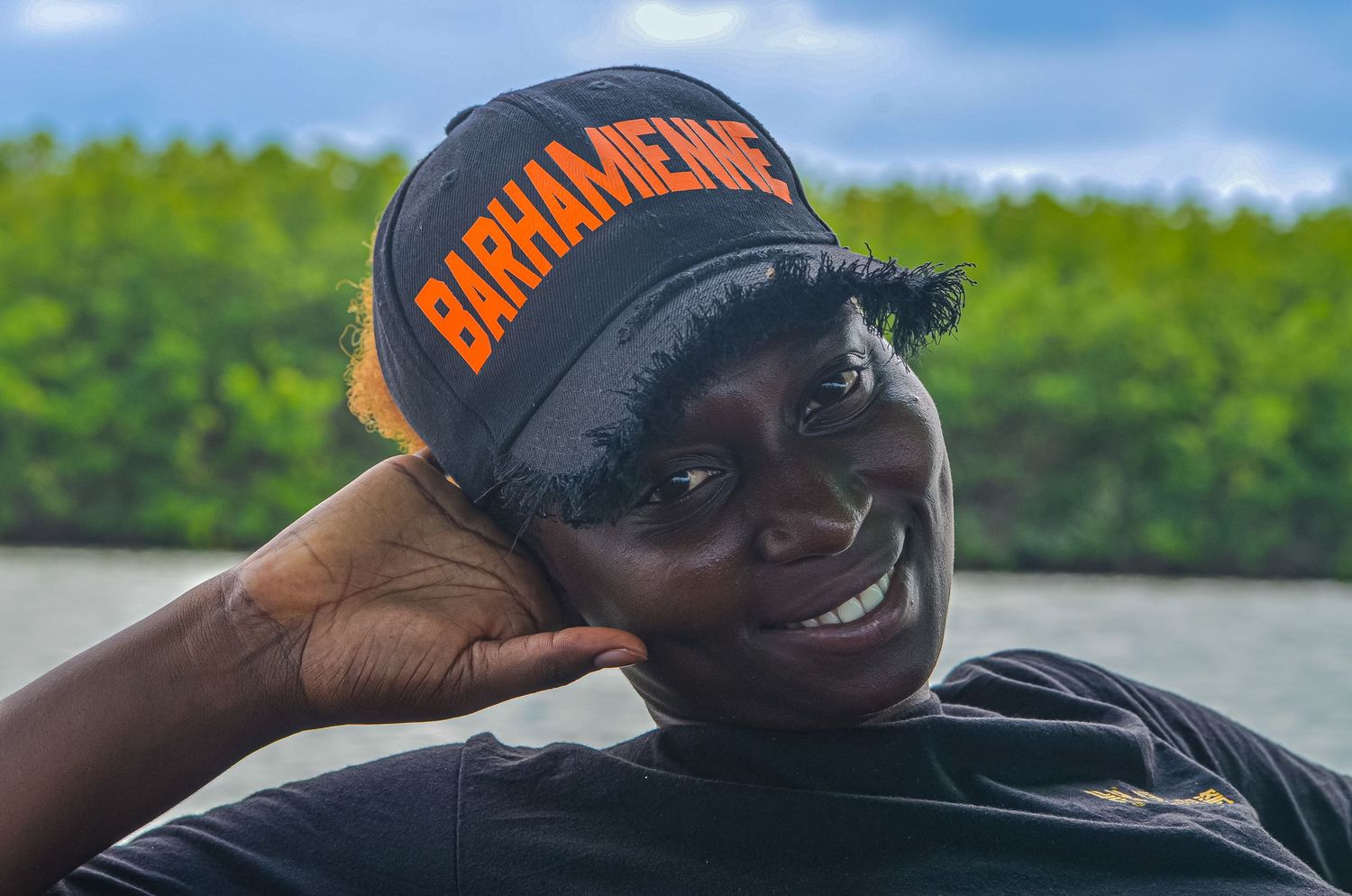 Smiling woman wearing a black Bahamienne cap and dark shirt outdoors with green foliage background.