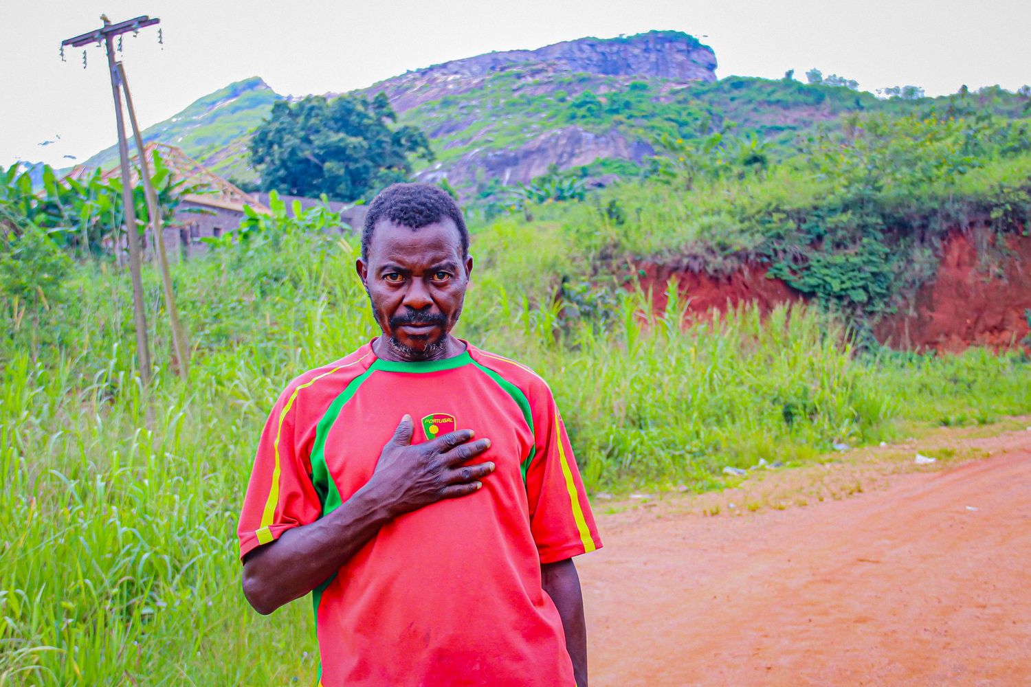 Smiling African man in a red and green soccer jersey standing on a dirt road with green hills behind him.