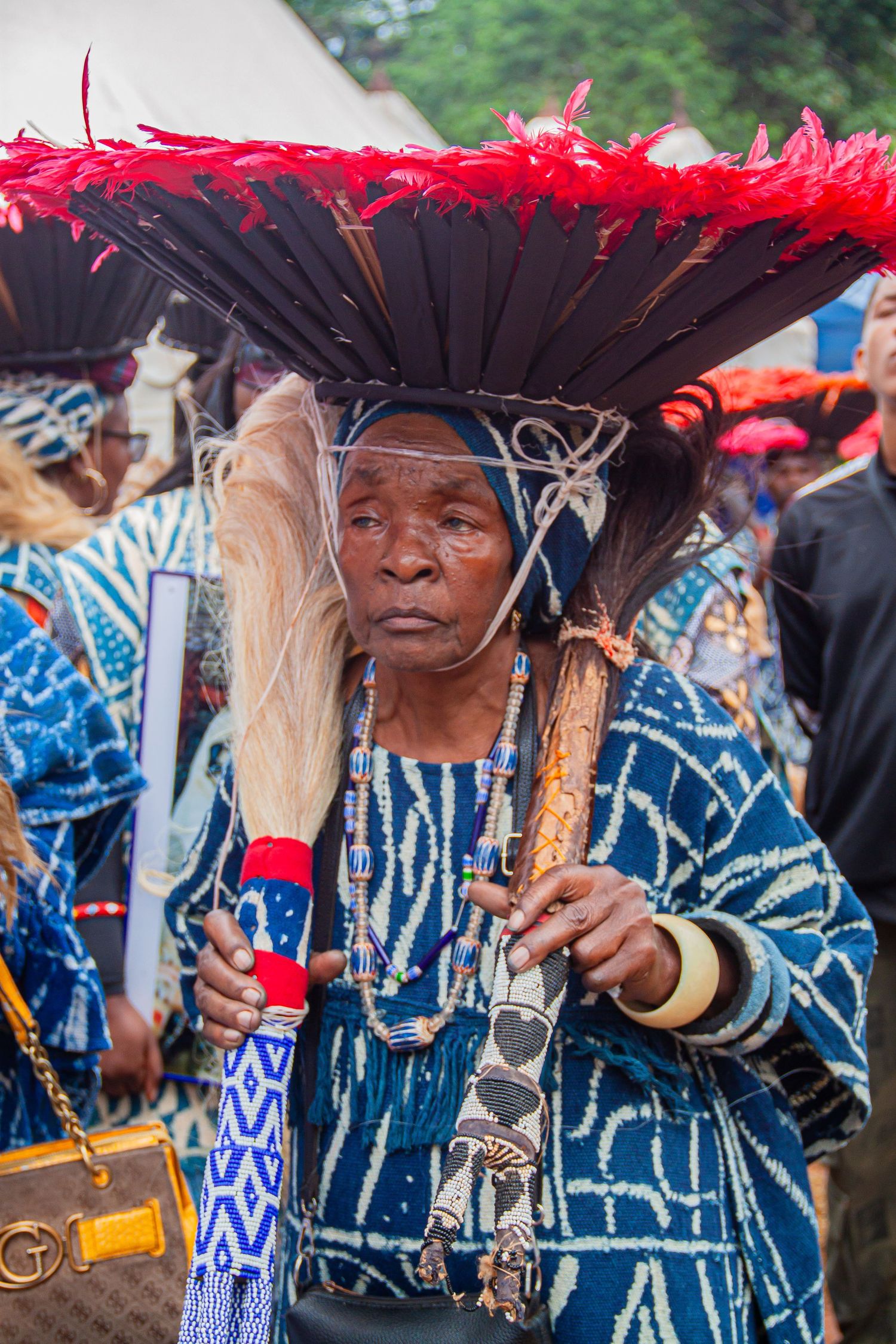 Elderly African woman in traditional blue batik attire with large feathered hat at cultural ceremony.