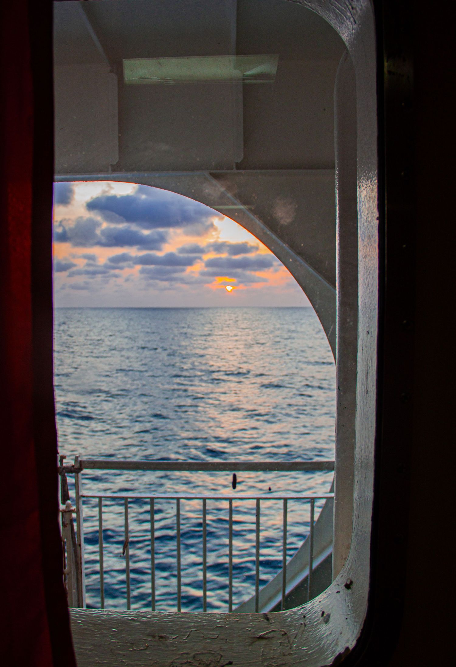 Ocean sunset viewed through an arched doorway on a ship balcony with railing and colorful sky.