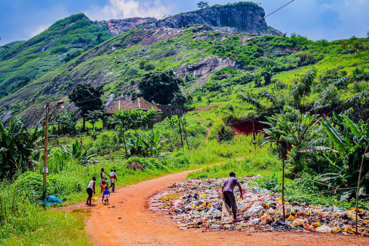 People walking along a red dirt road with a pile of garbage near a lush green mountain.